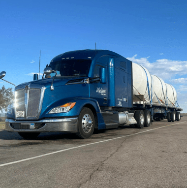 A blue Melton flatbed truck parked with an untarped load, secured with straps.